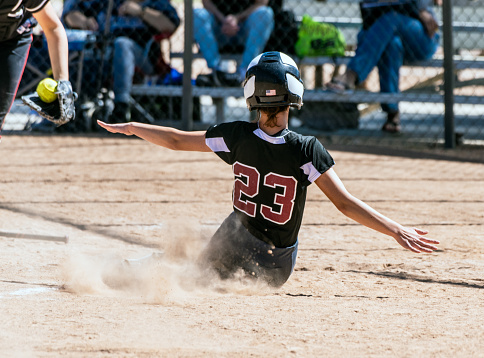 Female teenage softball player in black uniform sliding into home plate before the catcher can make the tag.