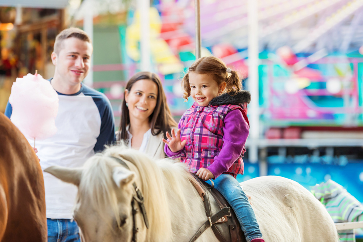 Girl enjoying pony ride, fun fair, parents watching her
