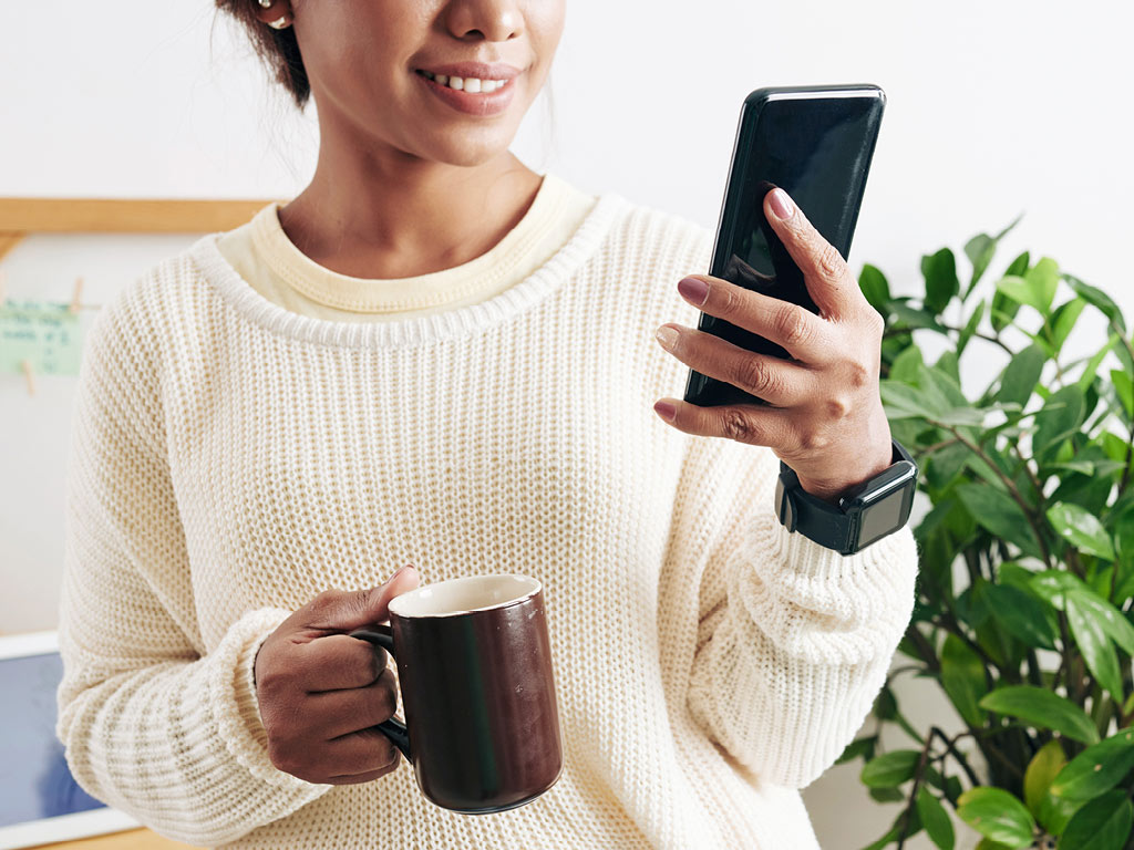 Woman looking at smartphone screen while holding mug of coffee in other hand.